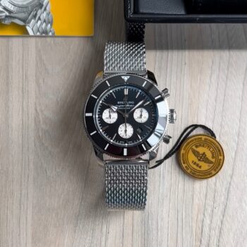 Super clone Breitling Superocean Heritage Chronograph 44 com mostrador preto, bezel cerâmico e pulseira milanese em aço sobre superfície de madeira clara ao lado da tag original Breitling